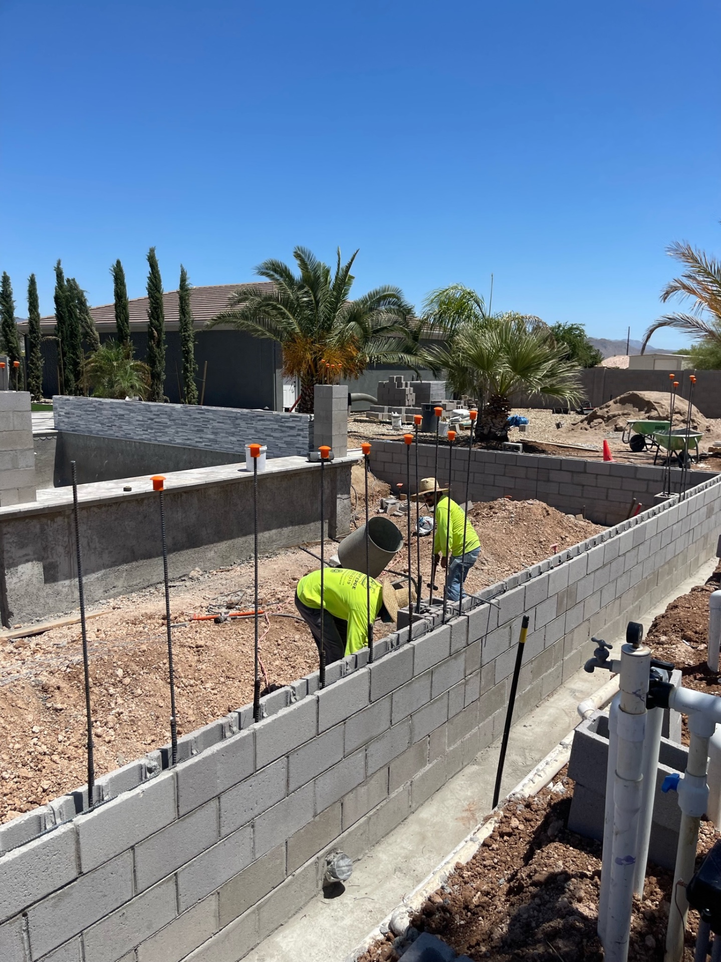 Crew members laying CMU block into a long backyard wall with vertical rebar and pool plumbing visible nearby