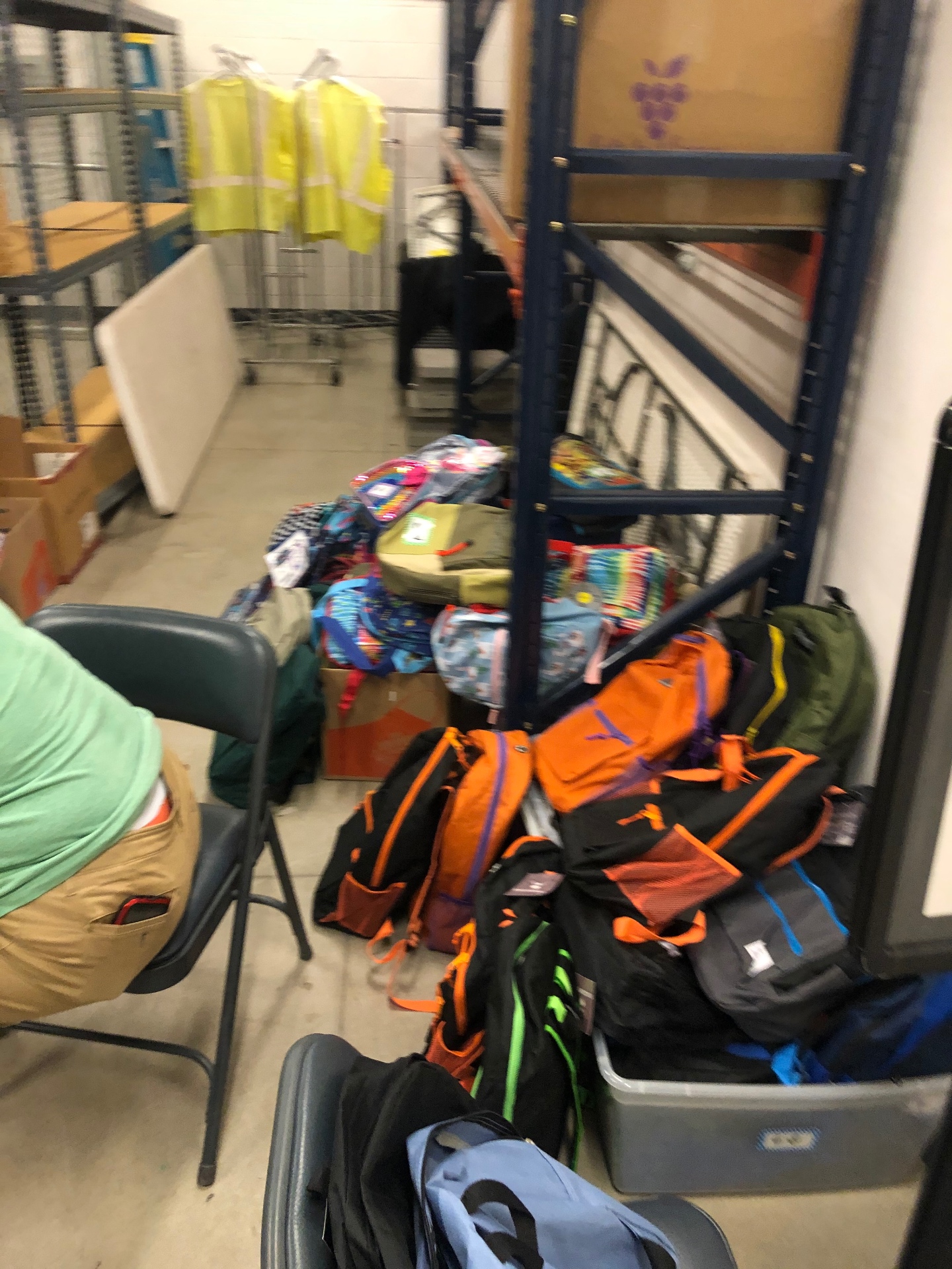 Volunteer in a hi-vis vest organizing piles of kid-pattern backpacks in the back warehouse