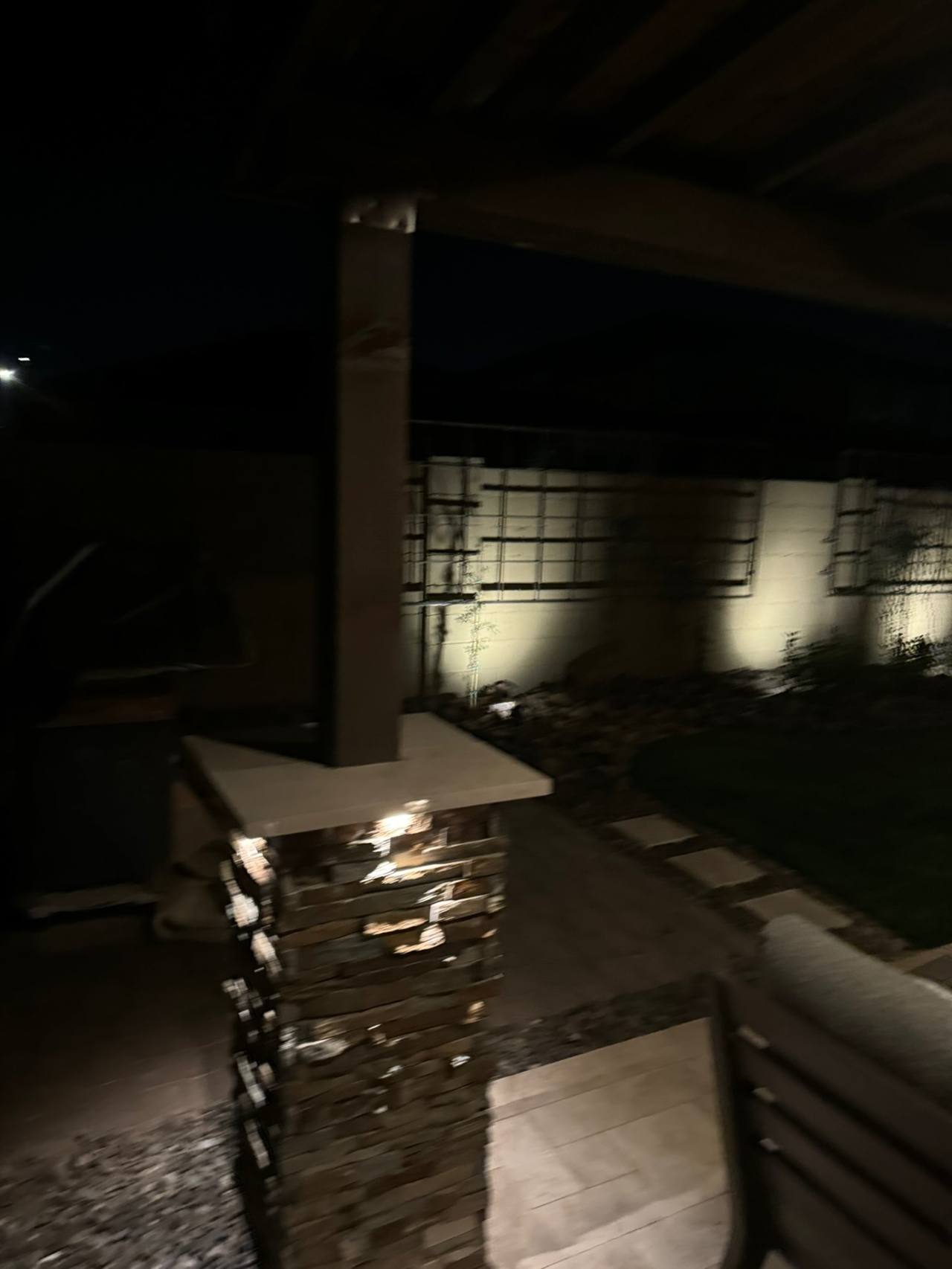 Covered-patio stacked-stone column, travertine cap, lit wall-wash fixtures, metal trellis panels, and paver-to-turf transition at night