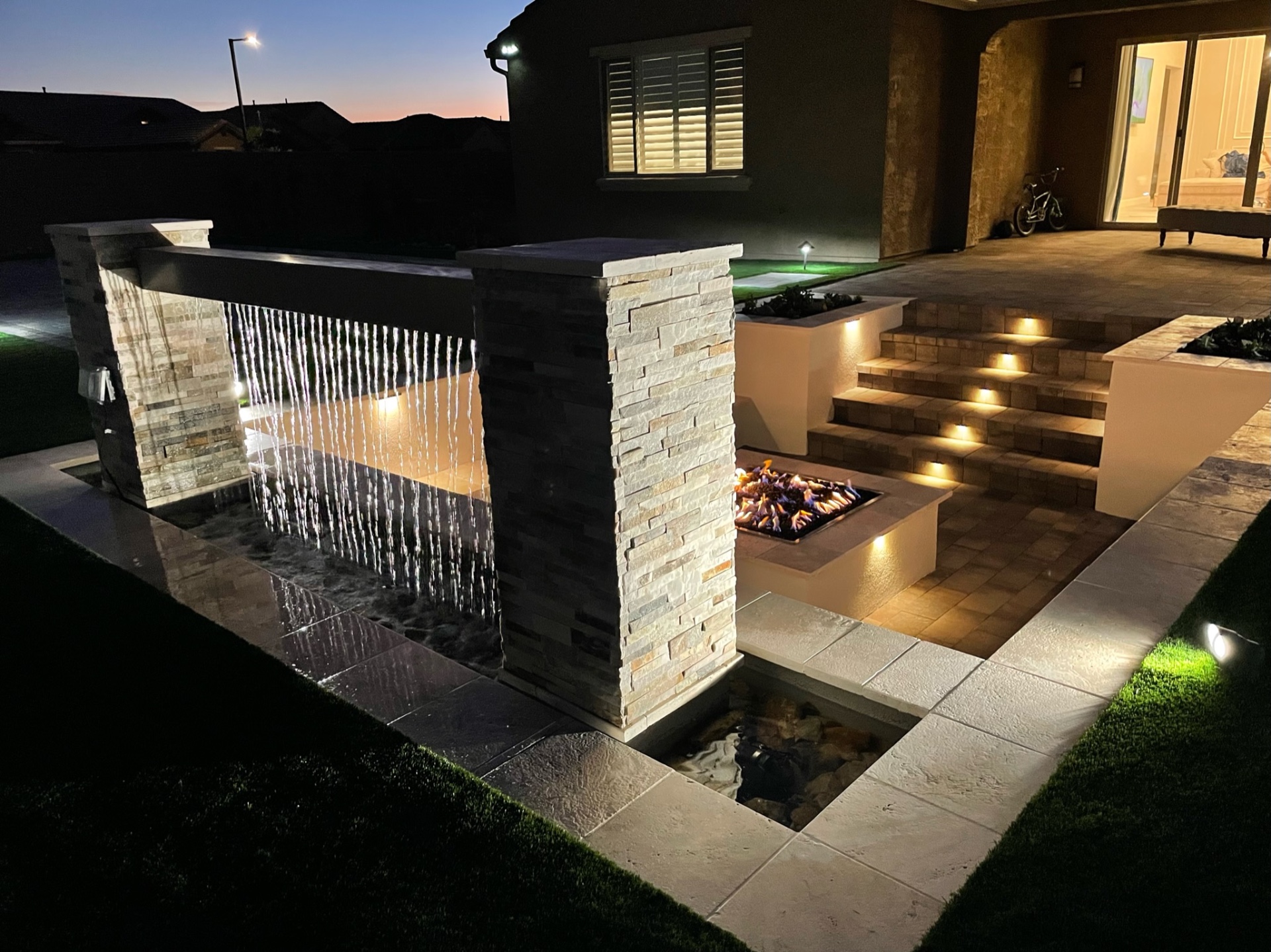Goodyear stacked-stone sheer-descent waterwall with paver step-down landing, gas firepit and lit paver stairs