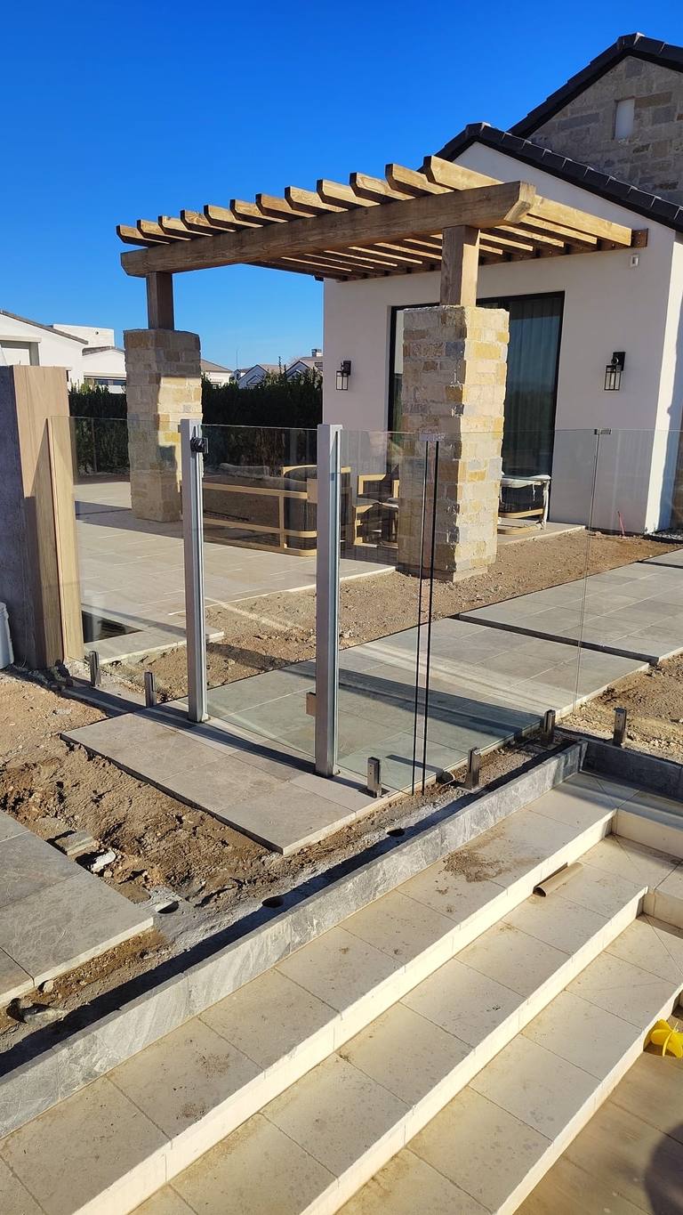 Surface-mount frameless glass fence with self-closing gate under a stone-and-timber pergola.