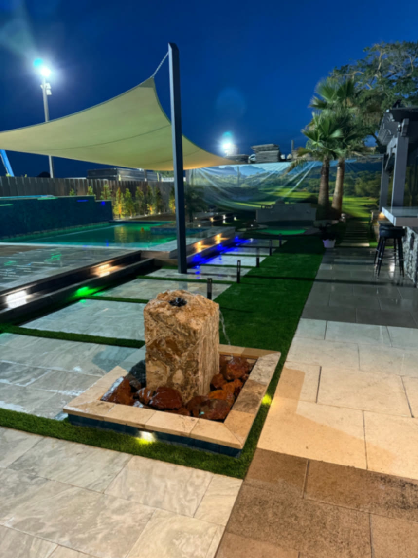Night view down the showroom yard with stone fountain feature, lit stepping pads, pool waterline tile, palm trees, and outdoor kitchen