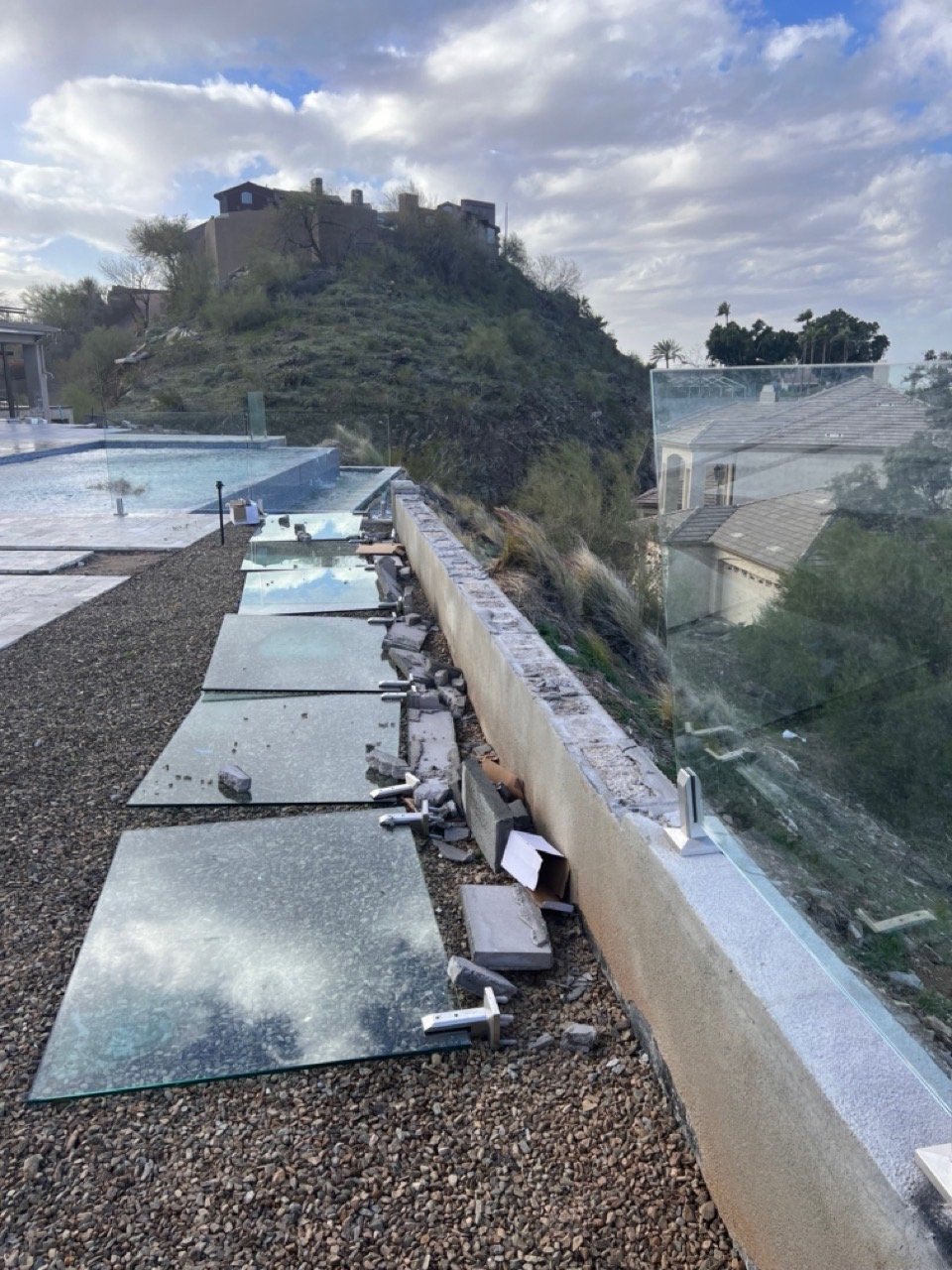 Failed glass pool fence installation on a Phoenix hillside — tempered glass panels laid on the ground next to a crumbled stucco wall cap with exposed bolt-down spigots.
