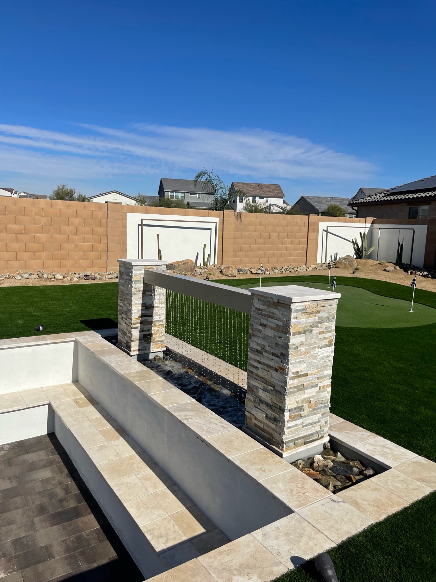 Goodyear stacked-stone rain-curtain water feature between travertine paver columns beside artificial turf lawn and putting green