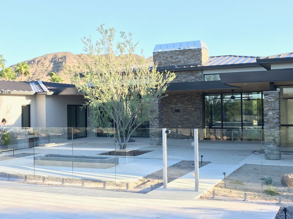 View across paver decking toward the home patio, outdoor living space, and frameless glass fence along the pool.