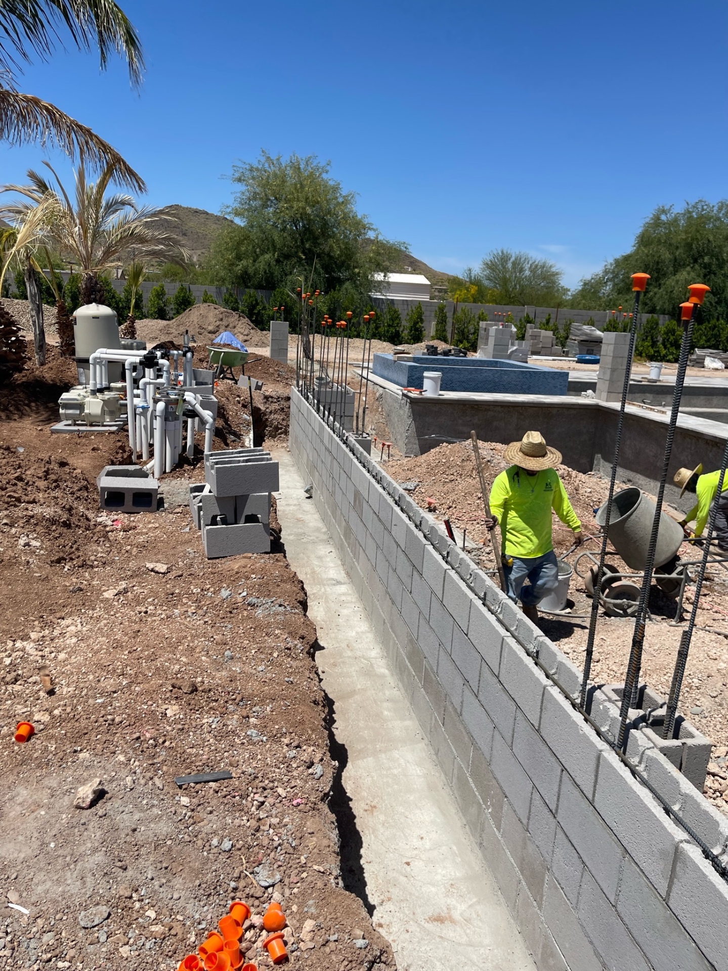 Crew member working a long block wall beside the pool equipment pad with raised blue-tiled spa, mountains, and palms behind