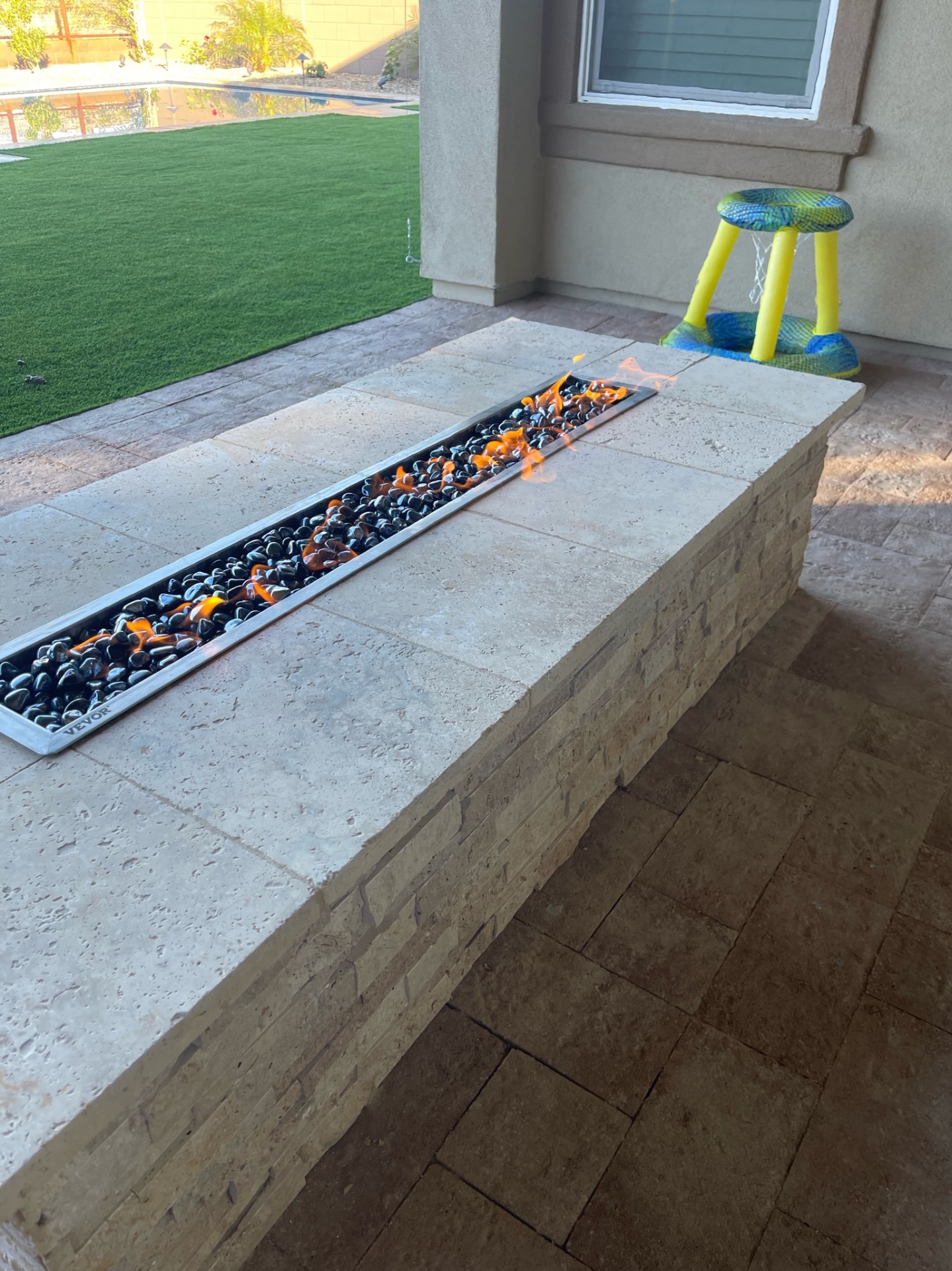 Peoria travertine-clad linear fire feature with stacked-stone base, black lava glass and travertine paver patio overlooking artificial turf lawn and pool by AE Outdoor Living