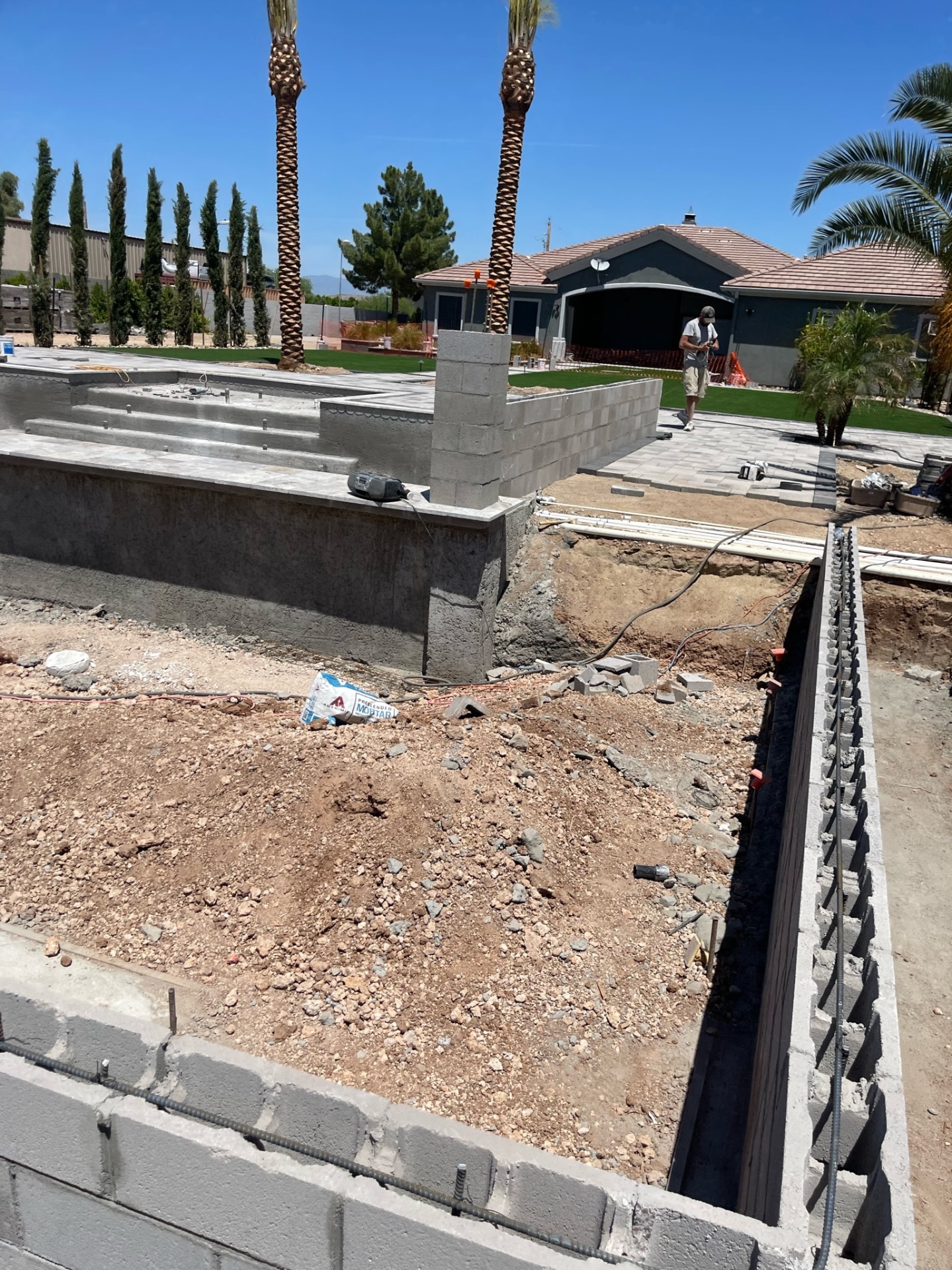 Mid-build view across the backyard with raised block walls, pool shell, paver patio, and front house visible past the construction zone