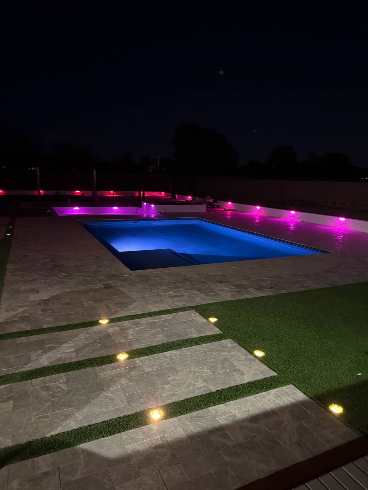 Rectangle pool and raised spa with pink LED wall lights, travertine paver deck, and turf ribbon steppers