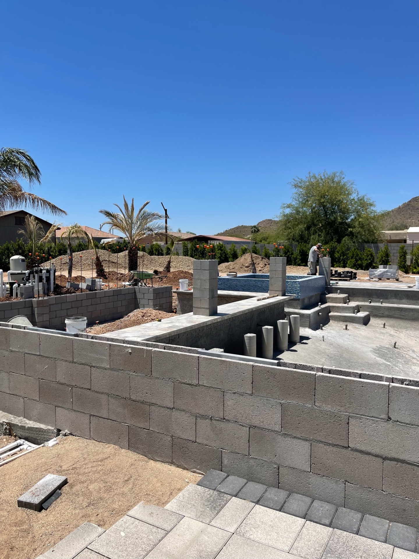 Wide view of block walls with raised blue-glass-tiled spa, pool shell, and equipment manifold under desert sky