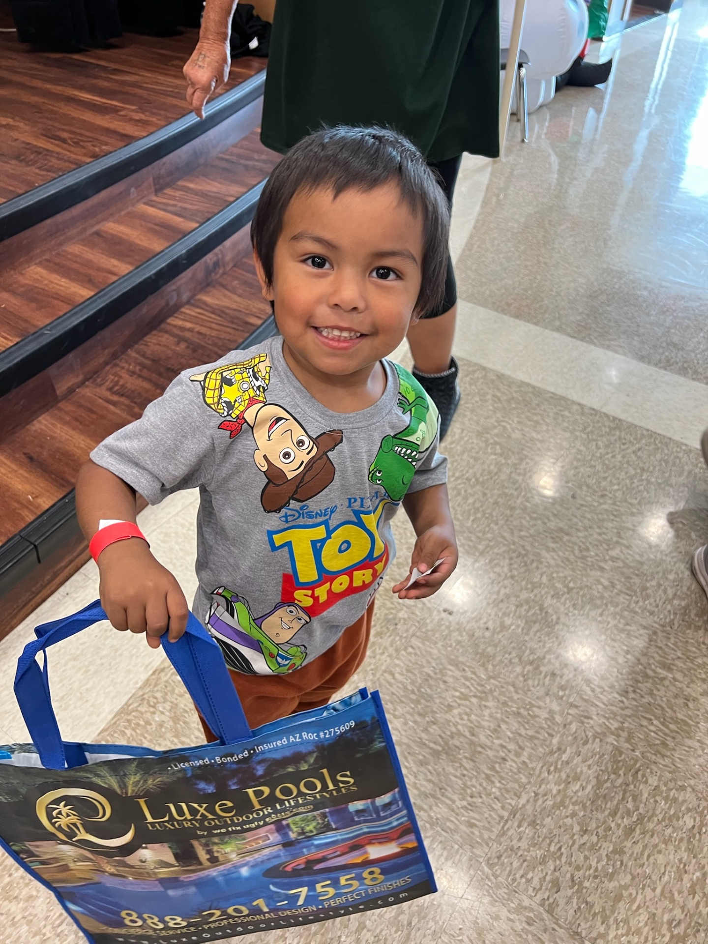 Young boy in a Toy Story shirt carrying an AE Outdoor Living Luxe Pools branded gift bag at a community toy distribution