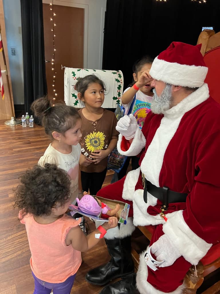 Santa kneeling and talking to a small group of young girls receiving toys at Shop With a Cop