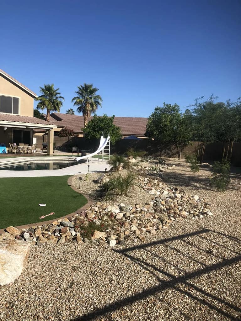 Rock-lined desert planting area with low-voltage landscape lights beside artificial turf and a backyard pool.