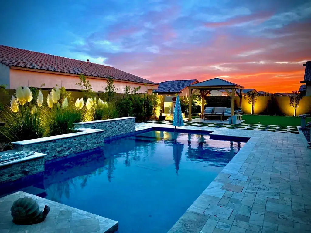 Completed AE Outdoor Living custom pool at sunset with lit water, travertine deck, gazebo and landscape lighting in the Phoenix metro