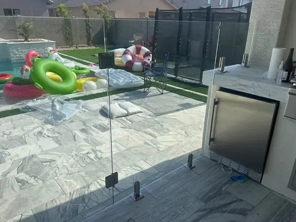 Frameless glass pool fence enclosing the pool deck and outdoor kitchen area with marble-look porcelain pavers.